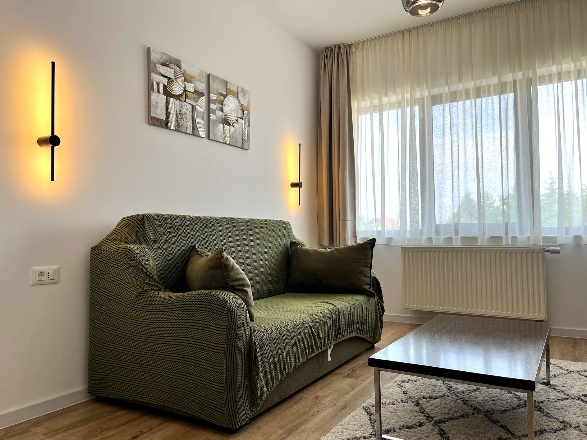 Canapea verde cu perne, două lămpi de perete, perdele deschise și tablouri abstracte într-un living luminos - apartament cu un dormitor in comuna Cristian, sat Cristian, Brasov, short-term rental