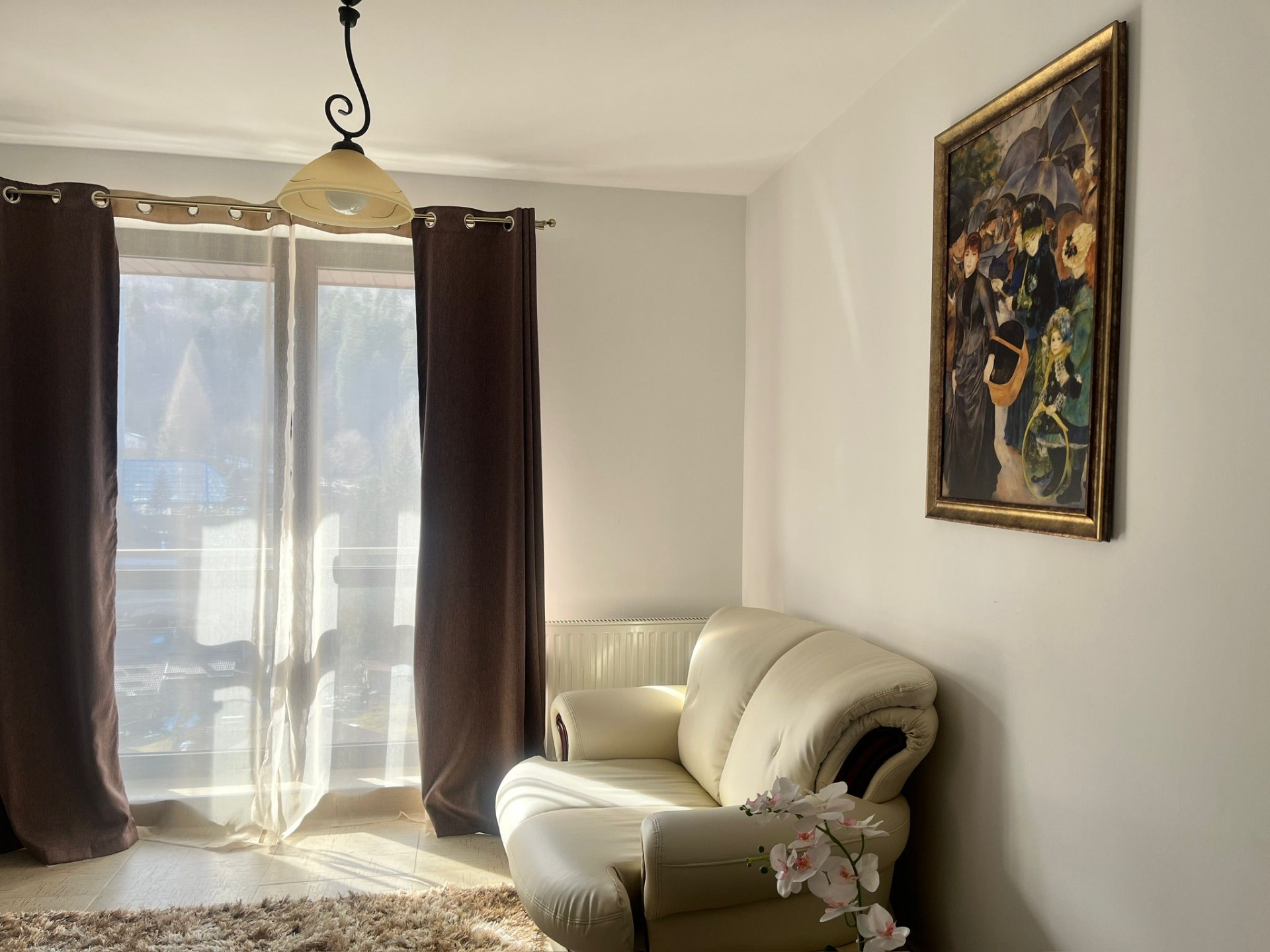 Apartament cu un dormitor, spatios si living room luminos cu draperii maro, fereastră, lampă suspendată și tablou încadrat pe perete; Sinaia, Colina Marei, parcare gratuita, short-term rental, cazare.