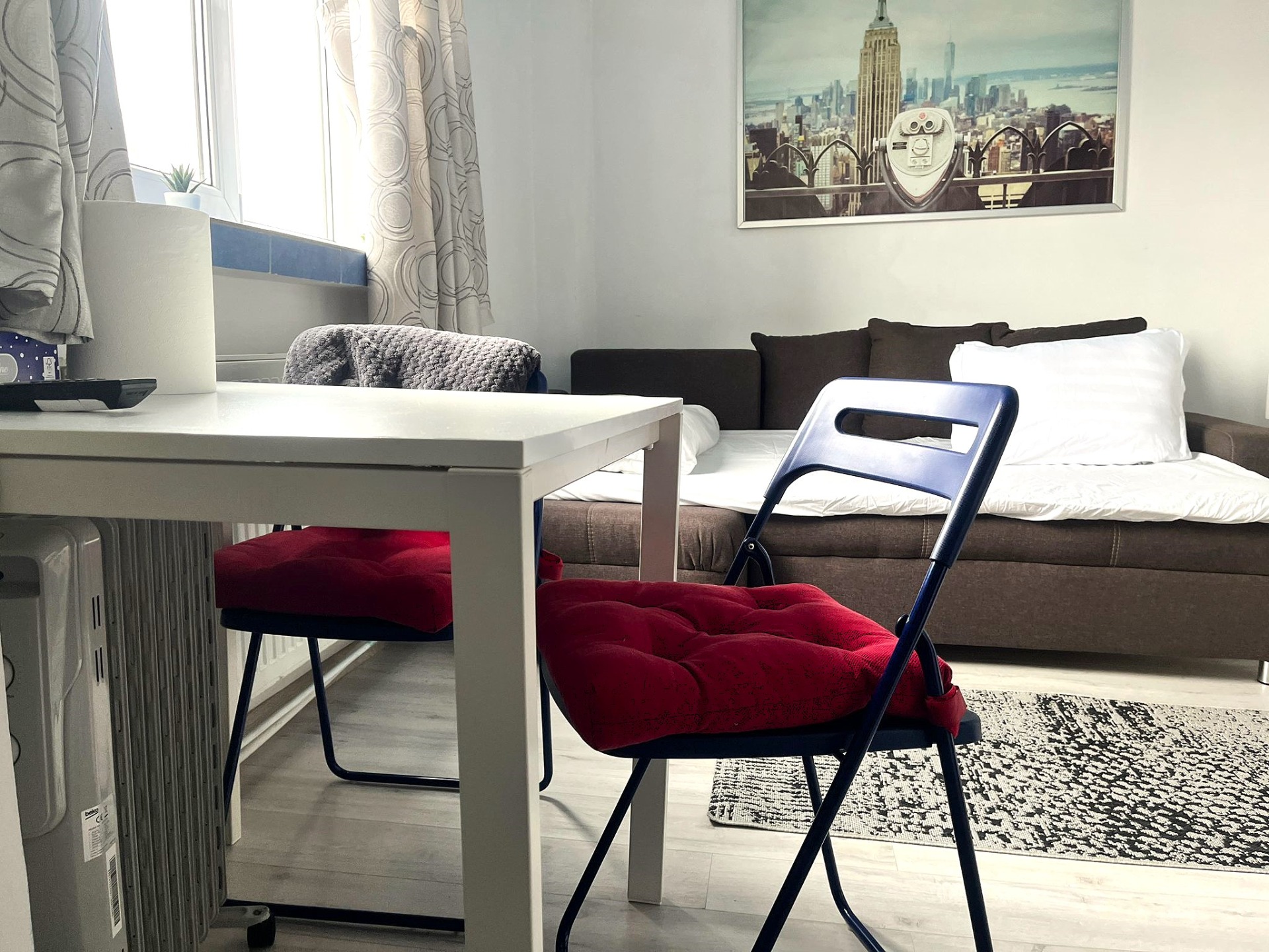 Studio in centrul Bucurestiului, Universitate; cazare termen scuze, cu canapea gri extensibila, masă albă, scaun albastru, pătură roșie și tablou cu skyline