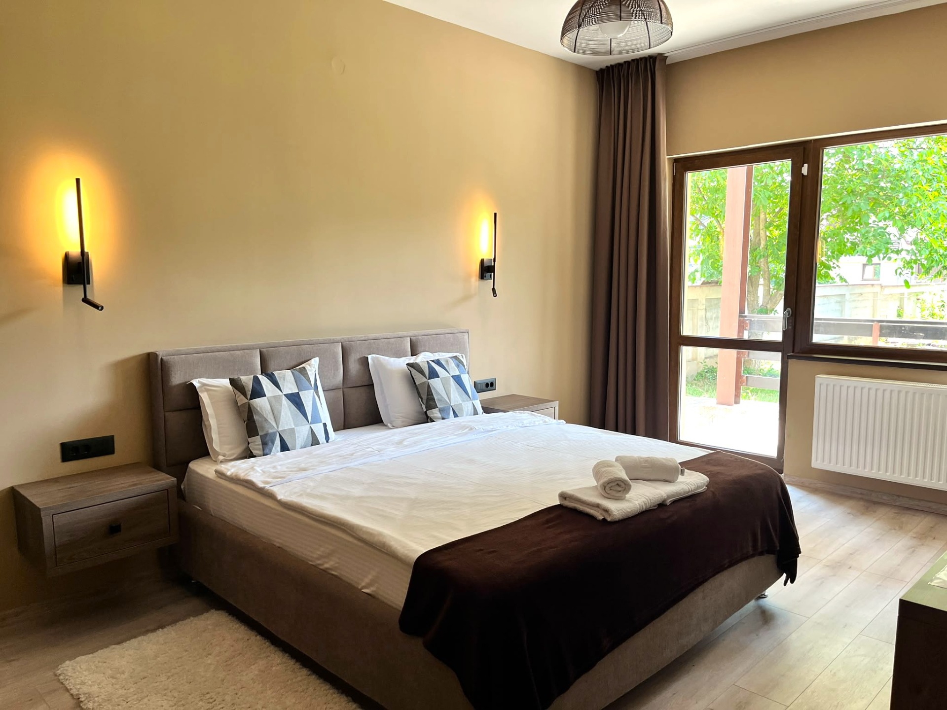 Dormitor modern cu pat dublu, perne geometrice, două lămpi de perete și ușă vitrată spre grădină; apartament cu doua dormitoare in Cristian, Brasov, cazare termen scurt